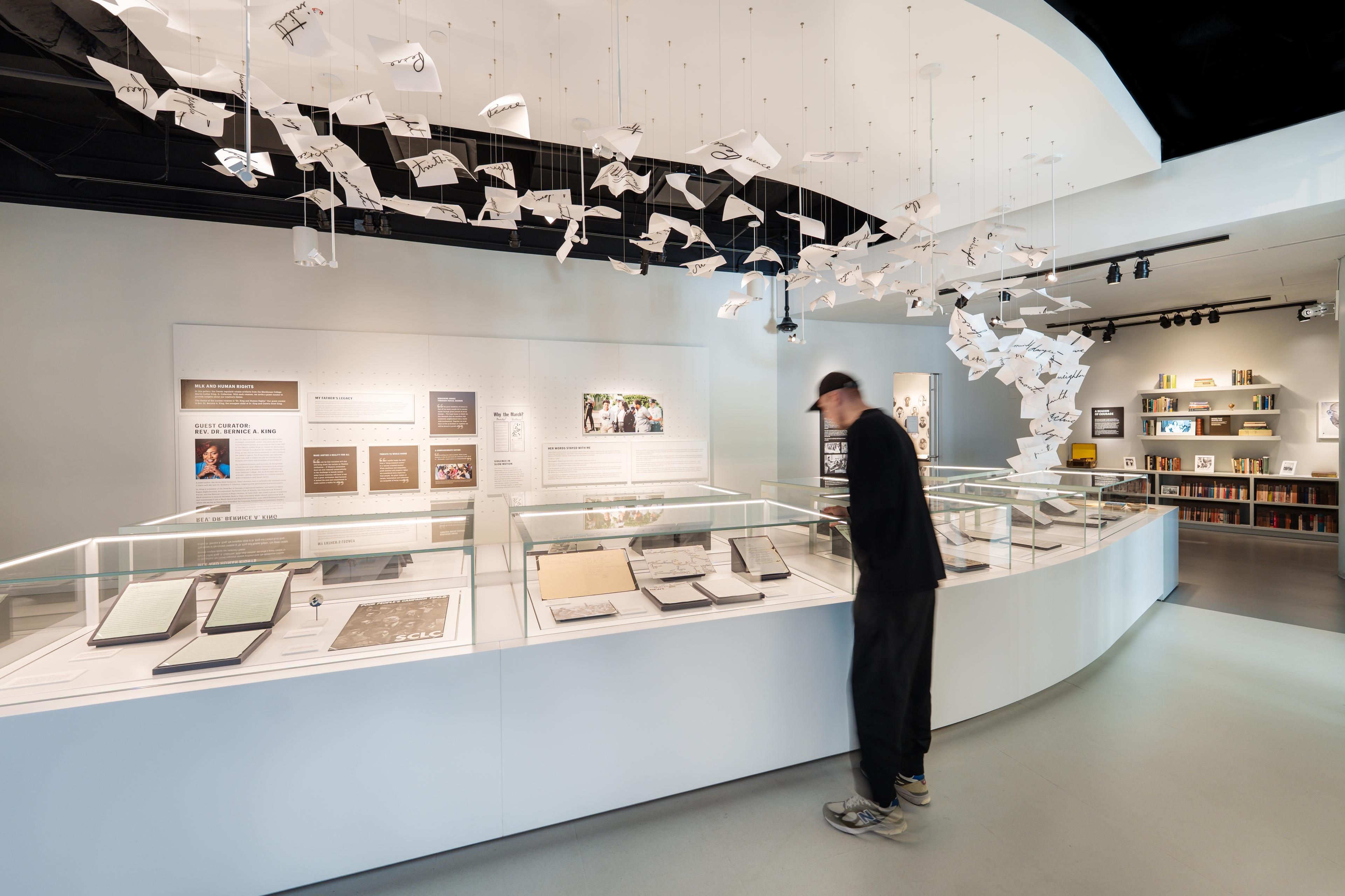 A museum gallery featuring archival materials related to Martin Luther King Jr. and the Civil Rights Movement. Curved display cases filled with historic documents are arranged beneath a dramatic ceiling installation of suspended handwritten pages that seem to rise and drift through the space. A visitor explores the exhibition, while interpretive displays and bookshelves in the background provide additional context